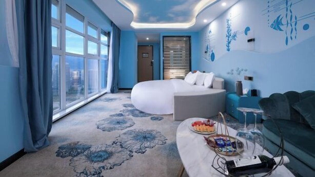 A modern hotel room with a large bed, a sofa, a dining table with snacks, and a view through large windows. - Hairui Hotel (Chongqing)