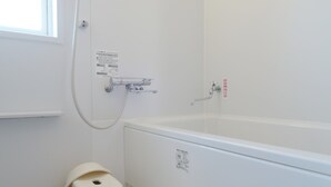 Une salle de bain équipée d’une baignoire, d’une douche et de toilettes. Une fenêtre laisse entrer la lumière naturelle.