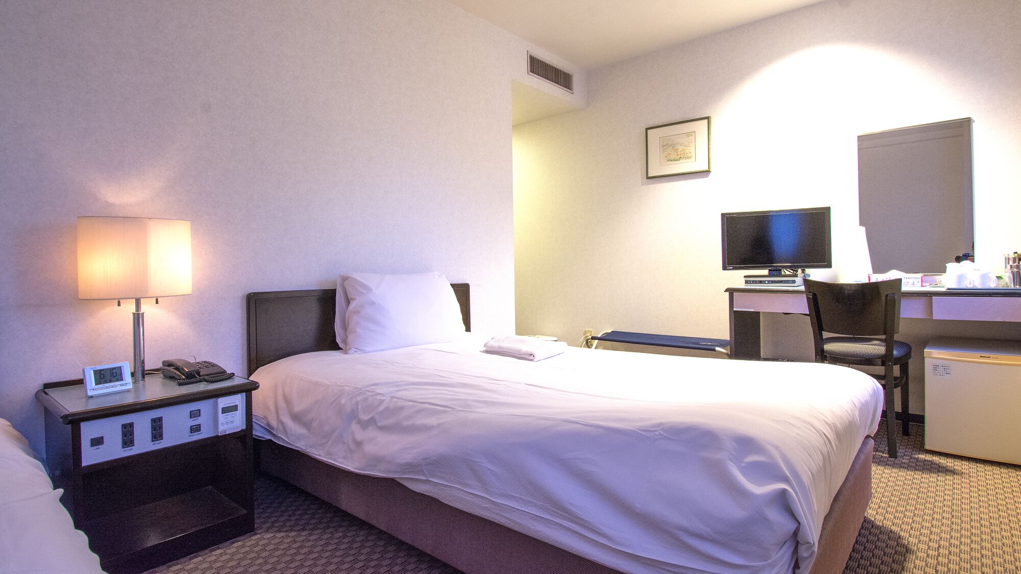 Une chambre d’hôtel comprenant un lit, une table de chevet avec une lampe, un bureau avec un ordinateur, une chaise et un petit réfrigérateur.