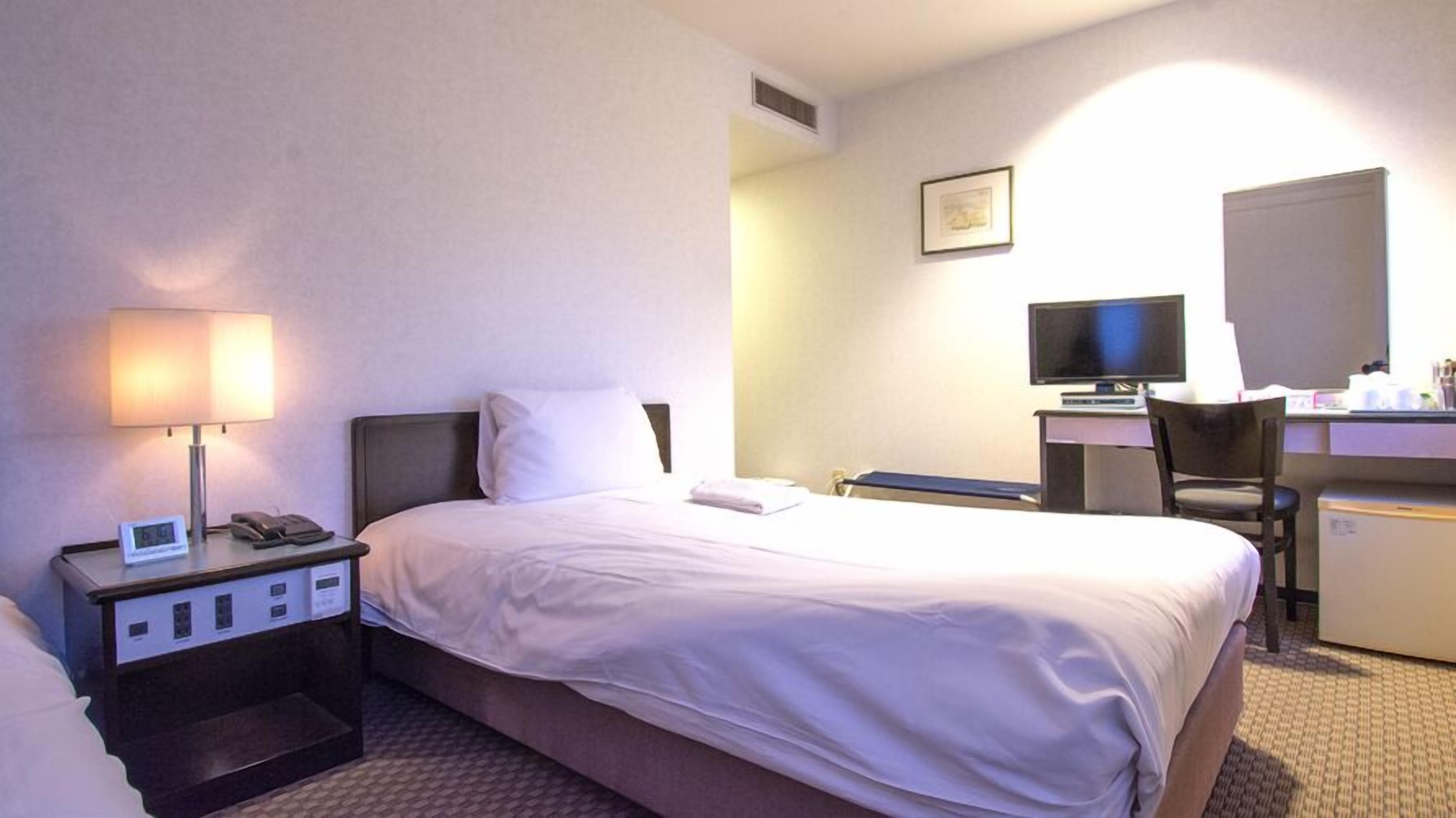 Ein Hotelzimmer mit Bett, Nachttisch mit Lampe, Schreibtisch mit Computer und Stuhl sowie einem kleinen Kühlschrank.