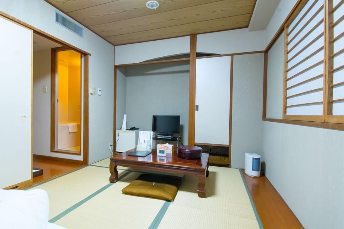Ein traditioneller japanischer Raum mit Tatami-Bodenbelag, einem Futon-Bett, einem Fernseher und einem Schreibtisch mit Computer und Telefon.
