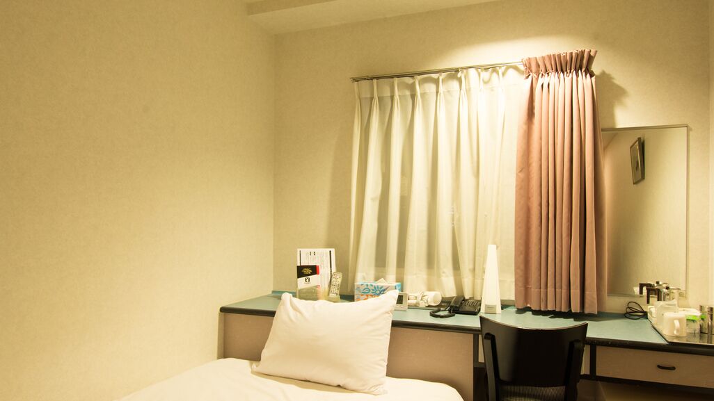 Ein Hotelzimmer mit Bett, Schreibtisch, Stuhl, Nachttisch, Lampe, Spiegel und einem Fenster mit Vorhängen.
