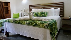 Una cama bien hecha con una colcha estampada con motivos tropicales, almohadas blancas y una toalla dispuesta en forma de corazón.