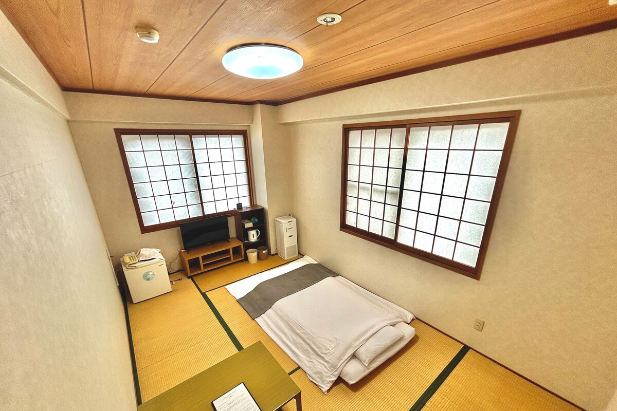 Une pièce de style japonais traditionnel, avec un sol recouvert de tatamis, un lit, un bureau, une télévision et un meuble.