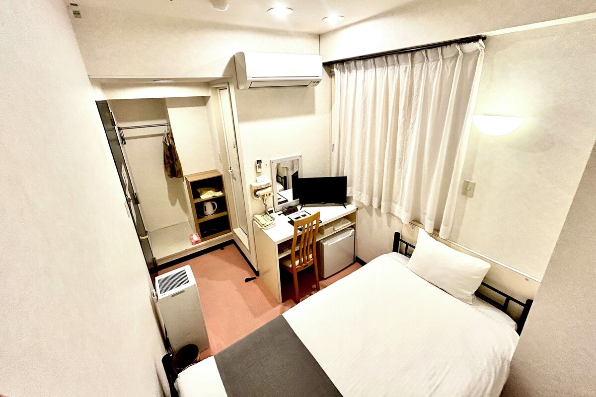 Une chambre d’hôtel avec un lit, un bureau avec un ordinateur, un miroir, un téléphone et une petite étagère avec une tasse.
