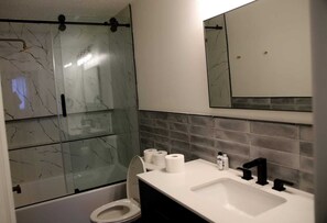 Ein modernes Badezimmer mit einer gläsernen Dusche, einem weißen Waschbecken mit schwarzen Wasserhähnen und einem Spiegel darüber.