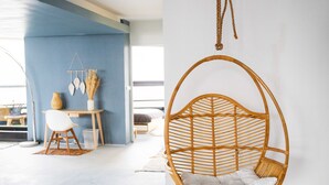 A room with a hanging rattan chair, a white chair, a wooden table, and a vase with dried plants.