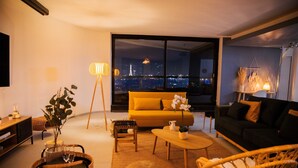 A modern living room with a yellow sofa, a black couch, wicker furniture, and a large window overlooking a cityscape at night.