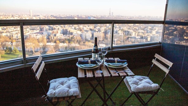A dining area with a table set for two, a bottle of wine, and two glasses. The table is on a balcony with a view of a cityscape.