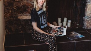 A person seated in front of a stone wall with a fireplace, holding a magazine.