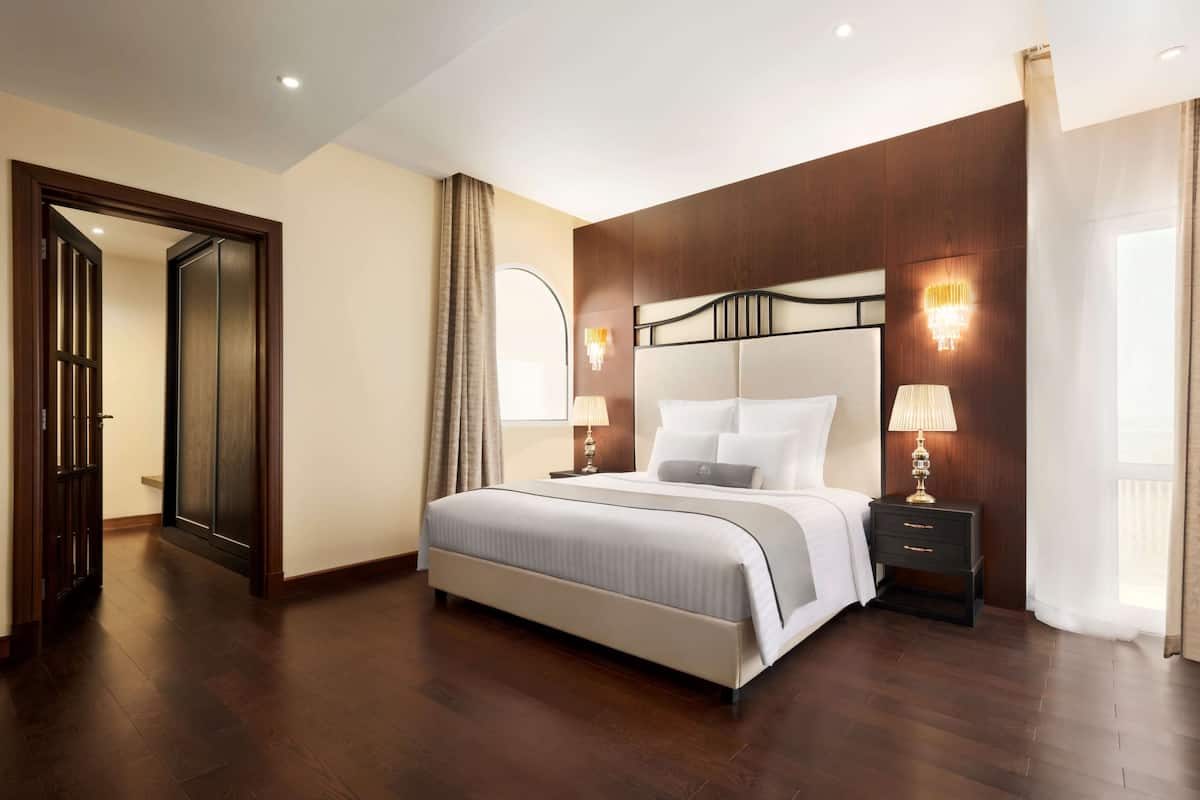Habitación de hotel con una cama grande, dos mesitas de noche con lámparas, un cabecero de madera y una puerta que da a otra habitación.