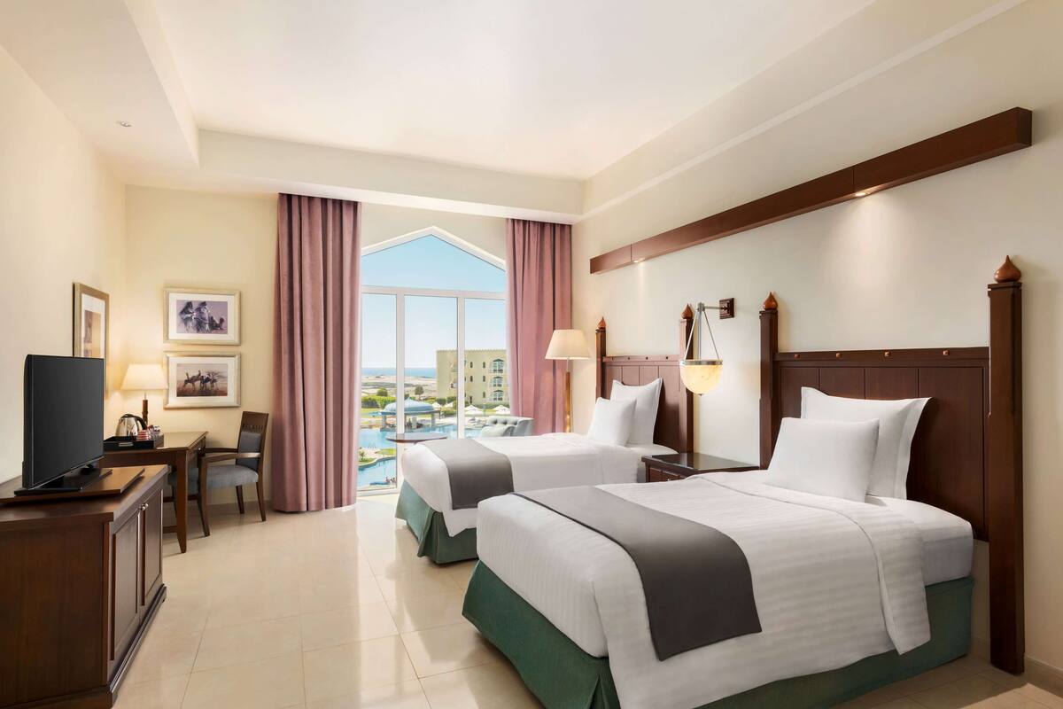 Habitación de hotel con dos camas, un escritorio con televisor, una silla y vista a una piscina y un edificio.