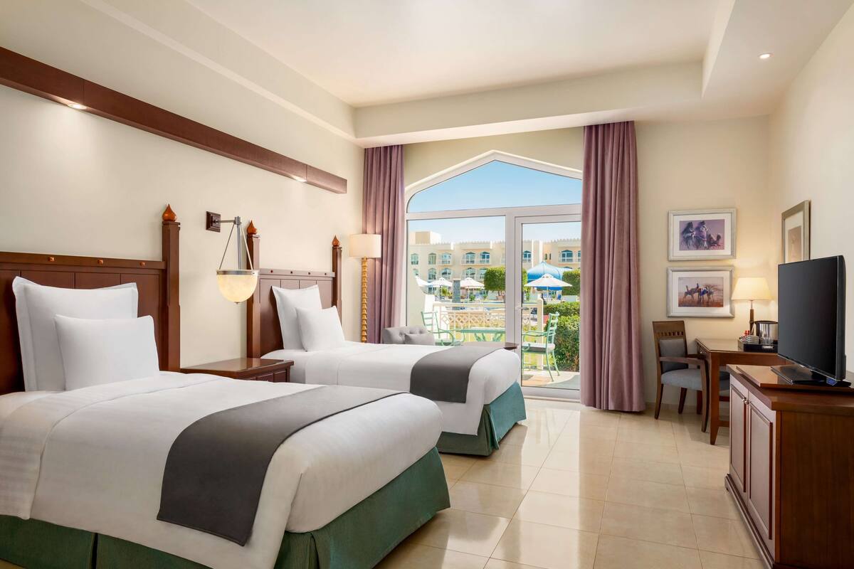 Habitación de hotel con dos camas, televisión, escritorio con silla y balcón con vista a edificios y zonas verdes.