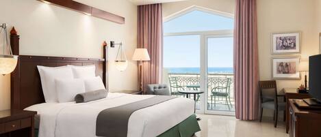 A hotel room with a large bed, a desk, a chair, a balcony with a view of the sea, and a TV.