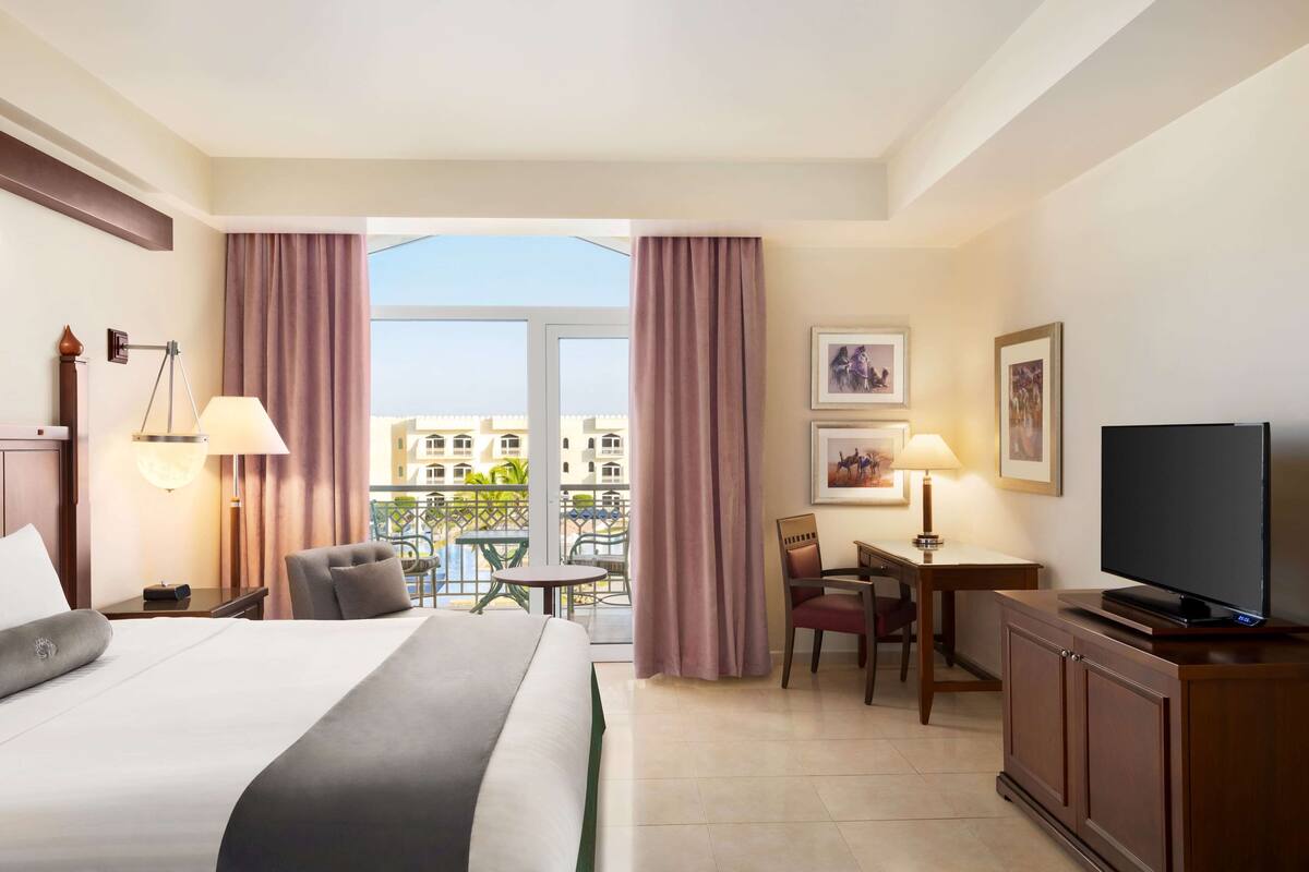 Habitación de hotel con cama, escritorio con silla, televisión y balcón con vista a edificios.