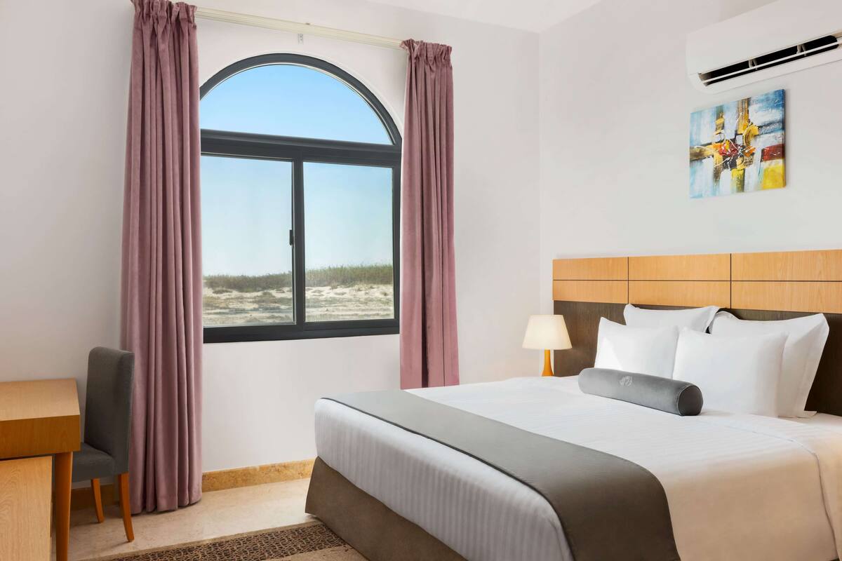 Una habitación de hotel con una cama, un escritorio, una silla, un ventanal con cortinas y un cuadro en la pared.
