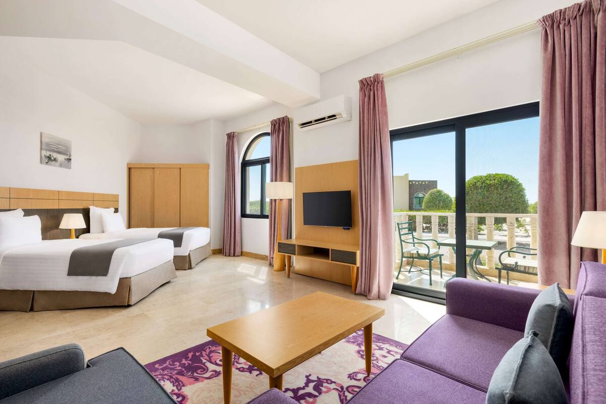 Habitación de hotel con cama, sofá, televisor y balcón con mobiliario exterior.