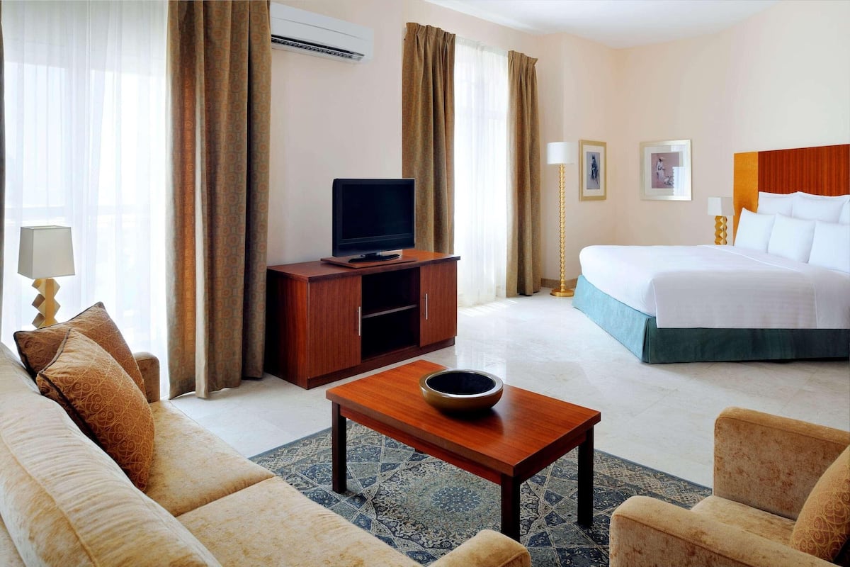 Habitación de hotel con una cama, un televisor sobre un soporte de madera, un sofá, una mesita de centro y una alfombra estampada.