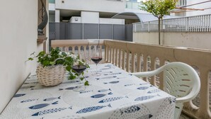 Un balcón con una mesa lista para dos personas, con un mantel estampado con motivos de pescado, una cesta de mimbre y una copa de vino tinto.