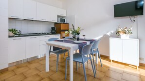 Eine moderne Küche mit weißen Schränken, einem Esstisch mit blauen Stühlen, einem an der Wand montierten Fernseher und einem kleinen Schrank mit Blumenarrangement.
