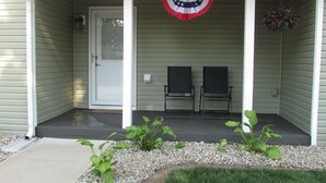 A covered porch with a door, two chairs, a small garden with pebbles and plants, and a hanging American flag. - 3 Bedroom House in Fort Recovery (Fort Recovery)