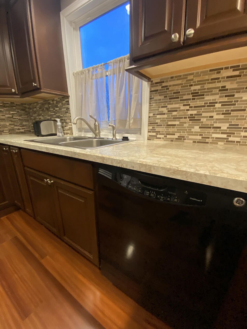 A kitchen with dark wooden cabinets, a built-in dishwasher, a window with a curtain, and a tiled backsplash.