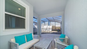 A covered outdoor seating area with white furniture and teal cushions, a large window with blinds, and a view of a greenhouse structure. - FA4381 Storey Lake Home 4Beds 3baths Private Pool and Free Waterpark (Kissimmee)
