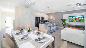 A modern kitchen with a central island, a dining area with a white table set, and a living area with a white sofa and a flat-screen TV. - FA4381 Storey Lake Home 4Beds 3baths Private Pool and Free Waterpark (Kissimmee)
