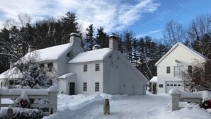Ein verschneiter Weg führt zu einem weißen Haus mit Schornstein, umgeben von Schnee und Bäumen.