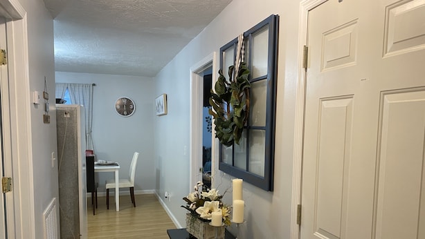 A hallway with a black table holding candles and a wreath, a white door, and a view into a dining area with a white chair and a clock on the wall. - Cozy Apartment (sleeps 6) (Kodak)