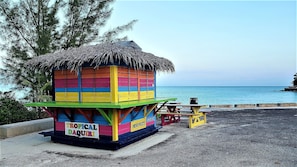 Uma cabana de praia colorida com telhado de palha, identificada como "Tropical Daquiri", e uma placa indicando que vende frutas frescas.