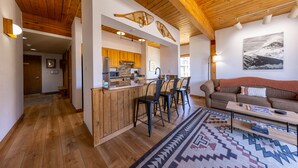 A spacious living area with a wooden ceiling, a kitchen with wooden cabinets, and a bar area with stools. - Wood Creek Condo | 2 Bedroom | Shared Hot Tub | Mtn Vew | Balcony | Parking (Mt. Crested Butte)