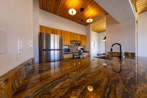 A modern kitchen with a granite countertop, stainless steel appliances, wooden cabinets, and a wooden ceiling.