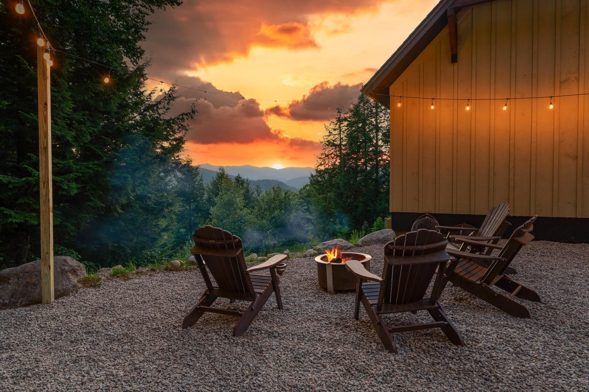 A patio with outdoor seating and a fire pit, overlooking a forested landscape with a sunset view.