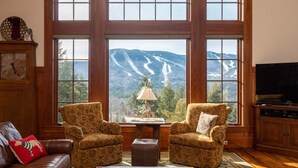 A cozy living room with large windows, two patterned armchairs, a wooden coffee table, a leather sofa, and a television.