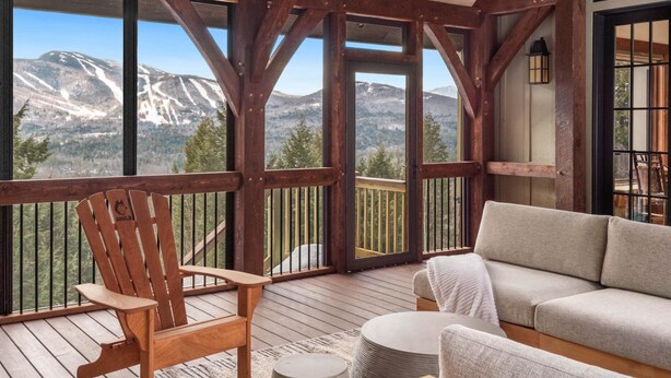 A wooden deck with a view of snow-capped mountains, a wooden Adirondack chair, and a gray sofa.
