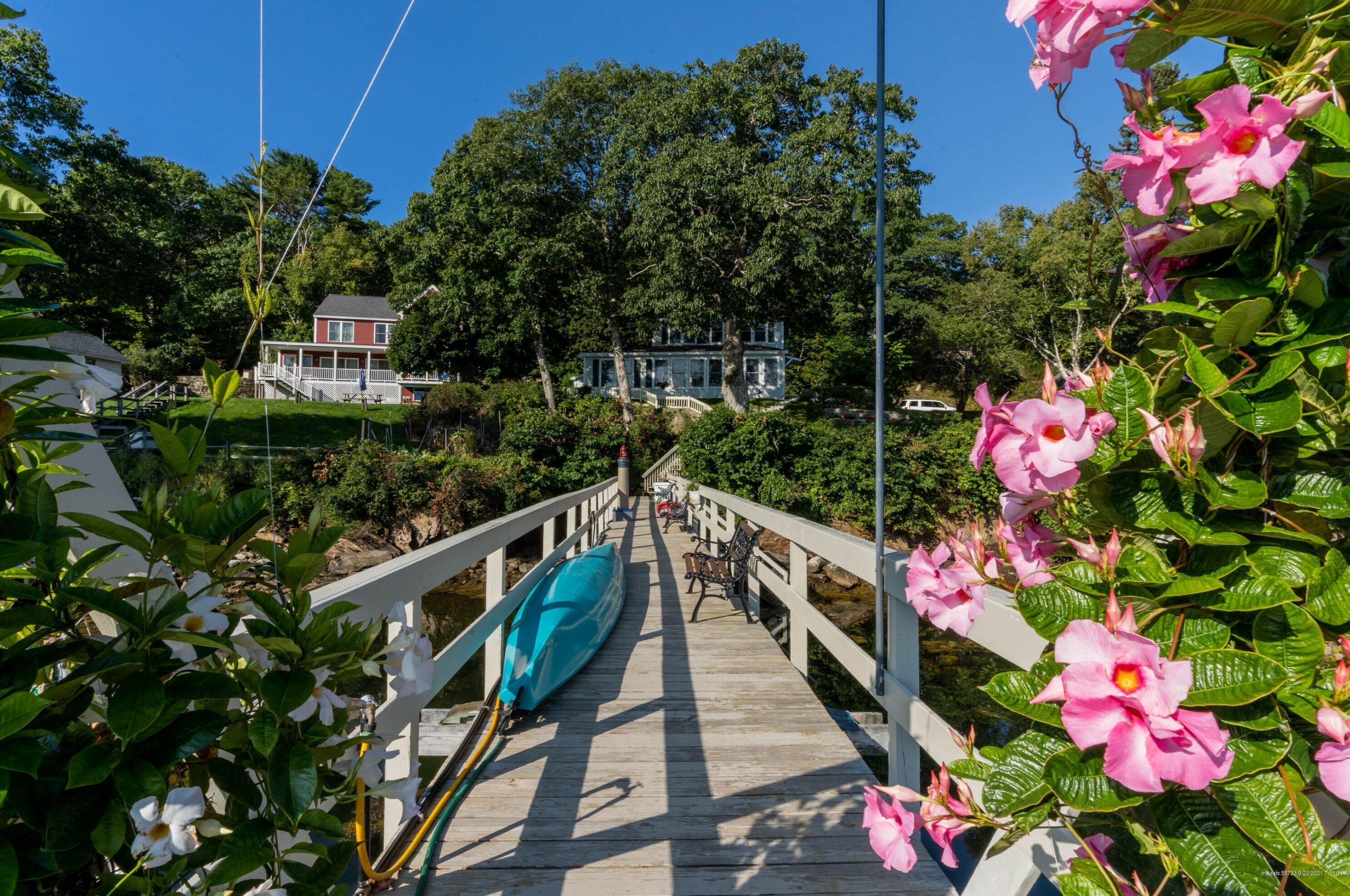 A wooden bridge with a kayak and benches, surrounded by lush greenery and a house in the background.
