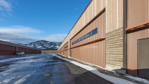 A large industrial building with multiple large doors and windows, a parking lot with snow, and a mountain in the background.