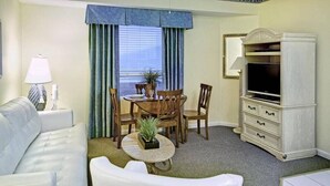 Habitación de hotel con un sofá blanco, una mesa de comedor con sillas, un mueble para televisión y vistas al exterior a través de una ventana con cortinas azules.