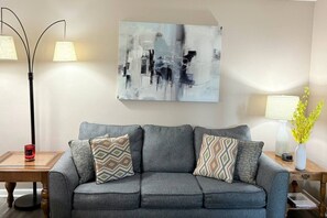 Ein Wohnzimmer mit grauem Sofa, gemusterten Kissen, einem modernen abstrakten Gemälde und zwei Stehlampen.