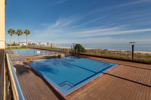 A swimming pool with a wooden deck, palm trees, and a view of the beach and cityscape. - Beachfront Penthouse with Wi-fi & Pool (San Javier, Murcia)