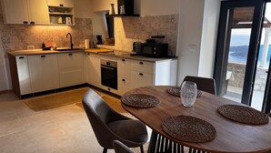 A modern kitchen with white cabinets, a wooden dining table, and a view of the sea through a glass door. - Cozy Coastal Retreat in Dubravica (Dubravica)