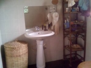 Un baño con un lavamanos blanco de pedestal, una cesta de mimbre y una estantería de madera con artículos de aseo.