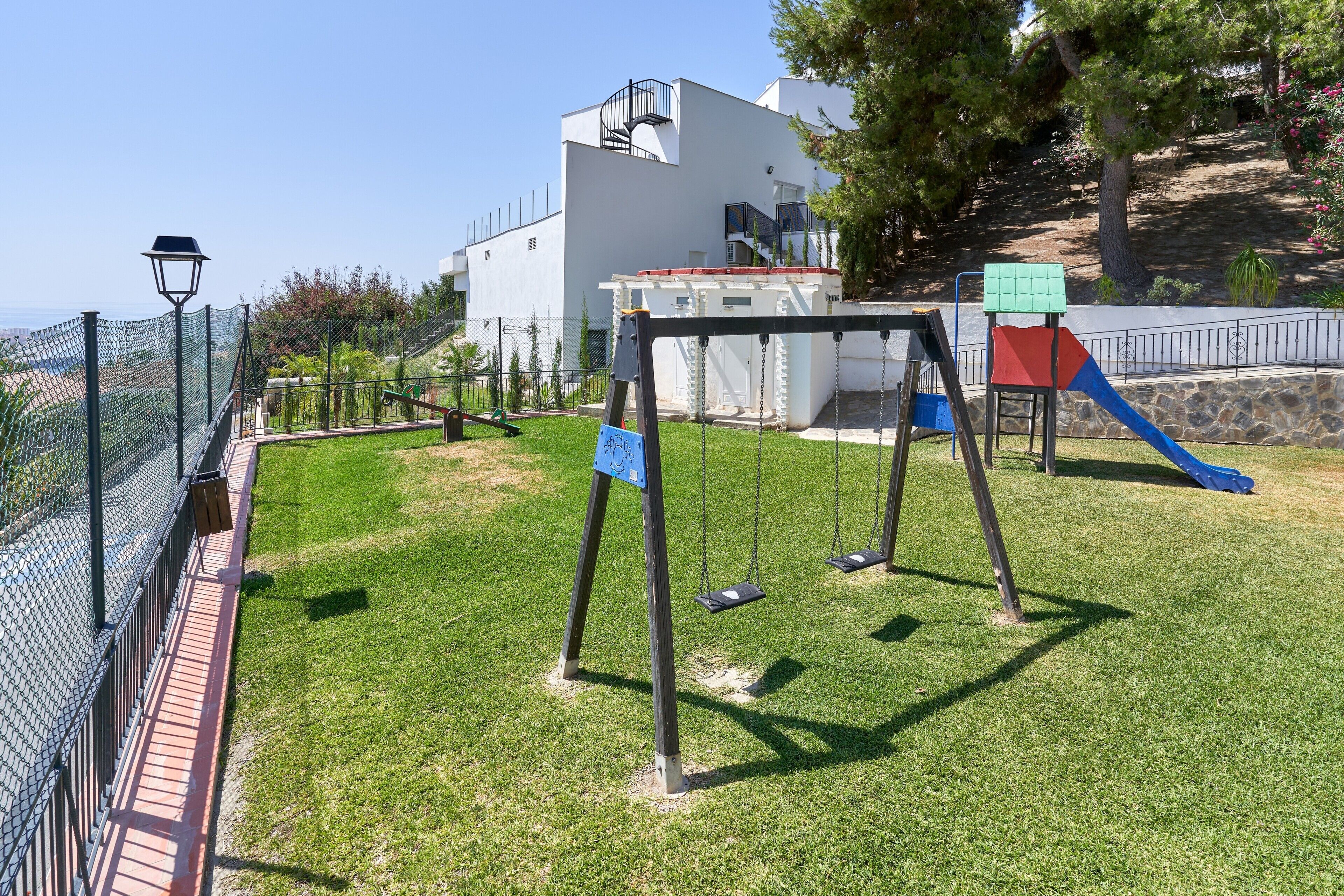 A playground with swings and a slide, surrounded by a fence and a building in the background.