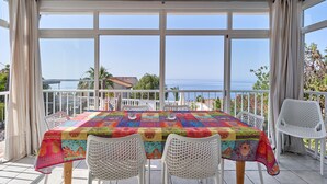 A covered outdoor dining area with a table and chairs, large windows, and a view of the sea.
