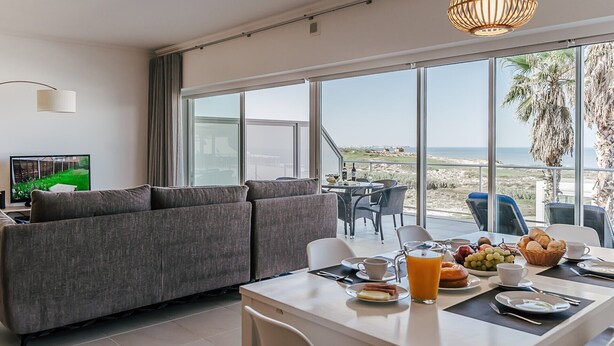 A modern living room with a large sofa, a dining area with breakfast items, and a view of the beach. - Apartment 'Beachfront Atlantic View A09' with Sea View and Wi-Fi (Óbidos)