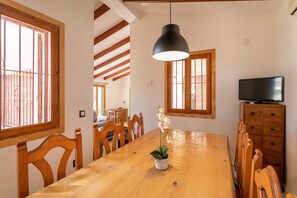 Ein Essbereich mit Holztisch, Stühlen, einer Topf-Orchidee, einer schwarzen Pendelleuchte und einem Fernseher auf einer Holzkommode.