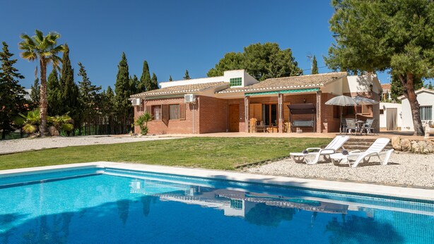 Vue sur une maison avec piscine, prise depuis le bord de la piscine, montrant un patio avec des chaises longues et un espace extérieur aménagé et couvert.