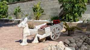 Un jardin agrémenté d’une statue d’âne décorative tirant une charrette, entouré de différentes plantes en pot et bordé d’un mur de pierre.
