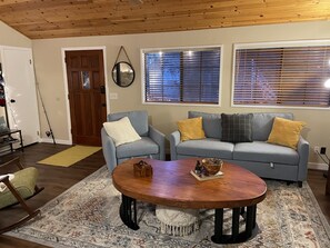 A living room with a wooden ceiling, two grey sofas with yellow and grey cushions, a wooden coffee table with decorative items, a patterned rug, and a wooden door. - 200,000 acre Los Padres National Forest as a back yard  (Frazier Park)